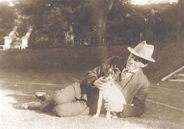 Man wearing suit and hat reclining on tennis court with dog, at 'St Brigids'. Woollahra Libraries Digital Archive, pf002458.